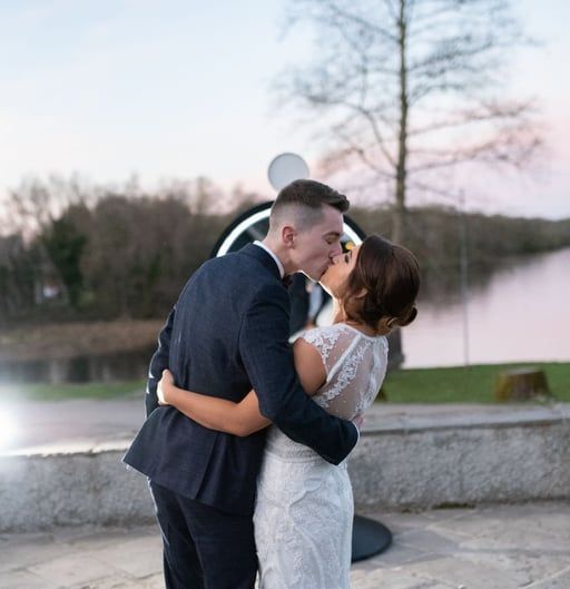 A couple kissing outdoors near a lake at dusk, standing before a circular light feature.
