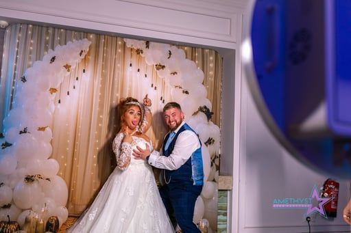 A couple in wedding attire poses in front of a white balloon arch and gold curtains.
