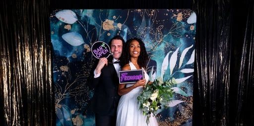 A couple poses in front of a blue and gold floral backdrop, holding signs that read 