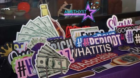 Assortment of colorful social media-themed photo booth props featuring money, liquor bottles, and hashtags on a purple cloth.