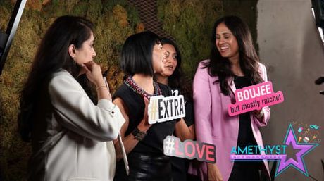 Four people laughing and posing with photo booth props against a green, moss-covered wall.