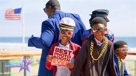 People wearing props and costumes at an outdoor photo booth with a flag and the ocean in the background.