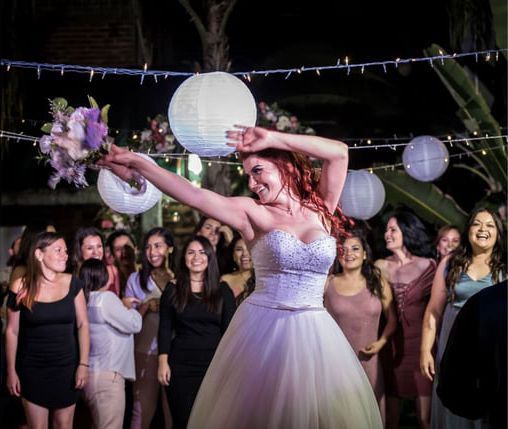 A bride in a white wedding gown joyfully tosses her bouquet to a crowd of guests at an evening outdoor reception.