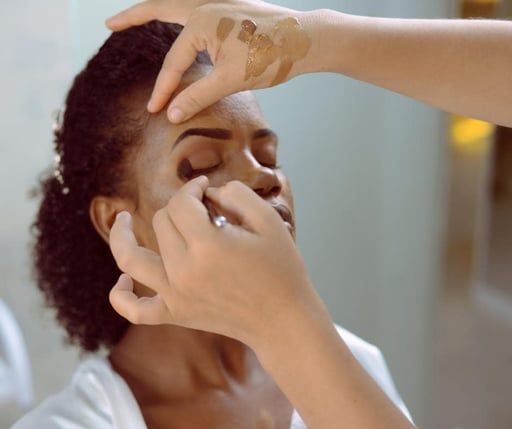 A makeup artist applies eyeshadow to a client’s eyelid using a brush, with foundation samples swatched on the artist’s hand.