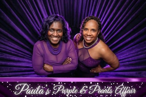 Two women in purple formal wear pose smiling against a pleated purple backdrop.