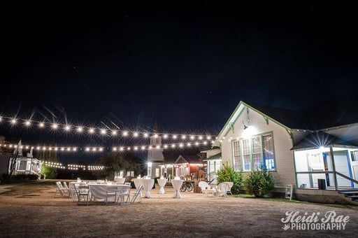 Outdoor evening scene with string lights above tables and chairs set up near a white cottage at a wedding venue.