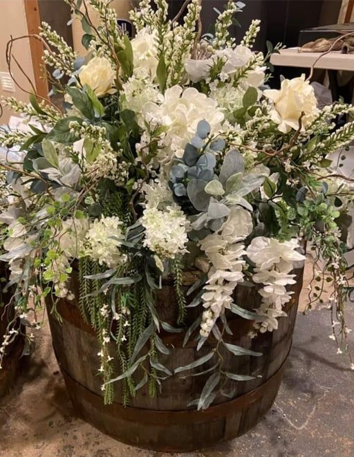 A lush, white floral arrangement featuring roses and greenery spilling out of a rustic wooden barrel.
