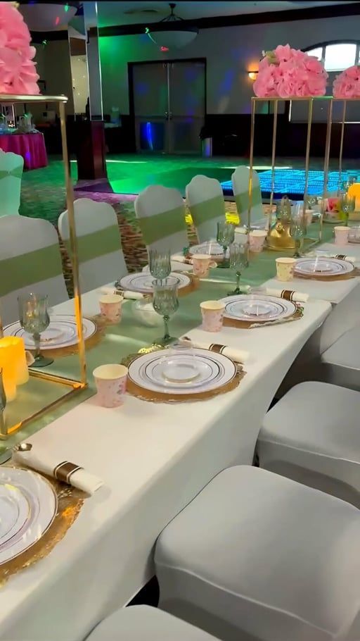 A long dining table set for an event with white tablecloths, grey chairs with green sashes, and floral centerpieces.