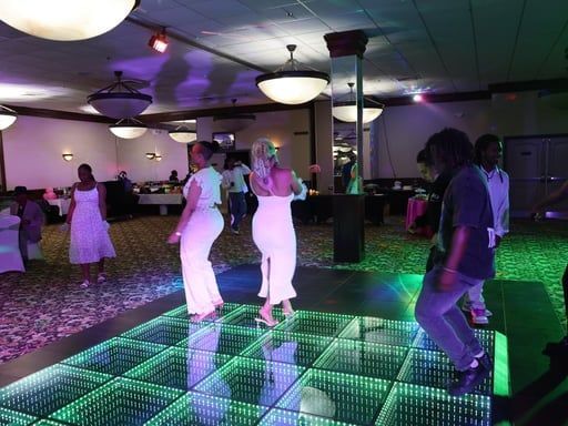 People dancing on a glowing LED dance floor in a ballroom decorated with pendant lights.