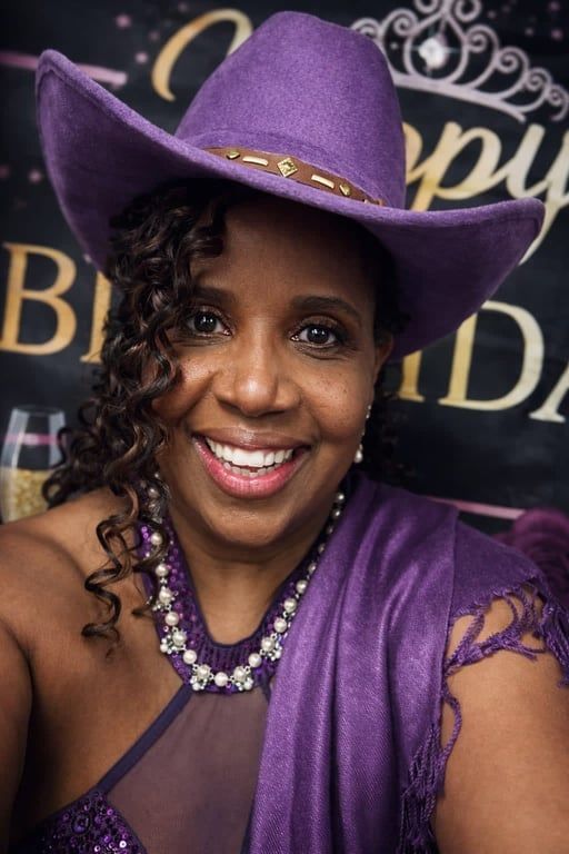 A smiling person wears a purple cowboy hat, a matching sequined top, and a pearl necklace against a birthday backdrop.