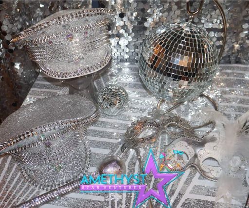 Two silver, rhinestone-encrusted captain's hats, a disco ball, and a masquerade mask on a shimmering, patterned surface.