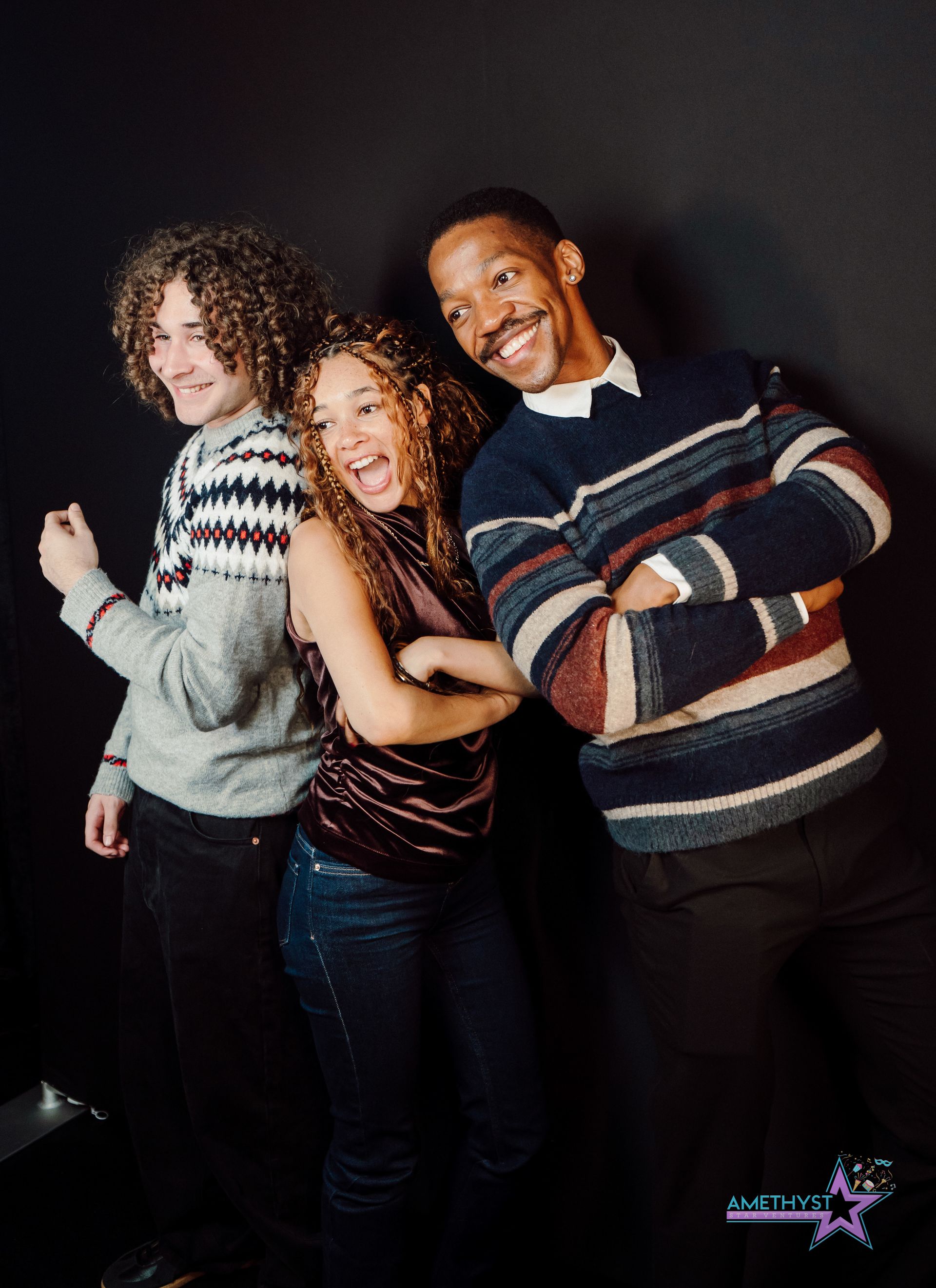 Three smiling people pose together against a dark background, wearing striped sweaters.