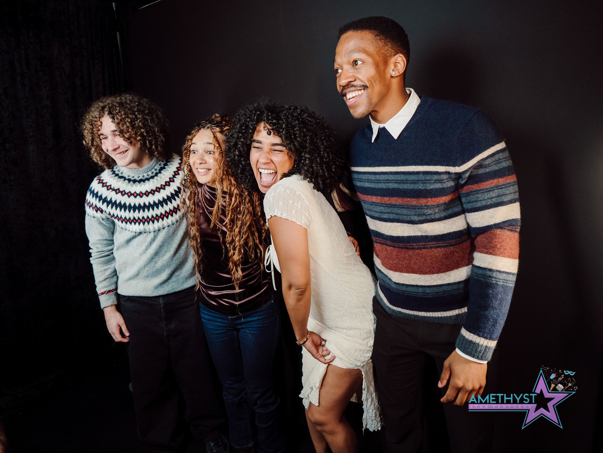 Four smiling people posing together against a dark background, wearing striped sweaters.