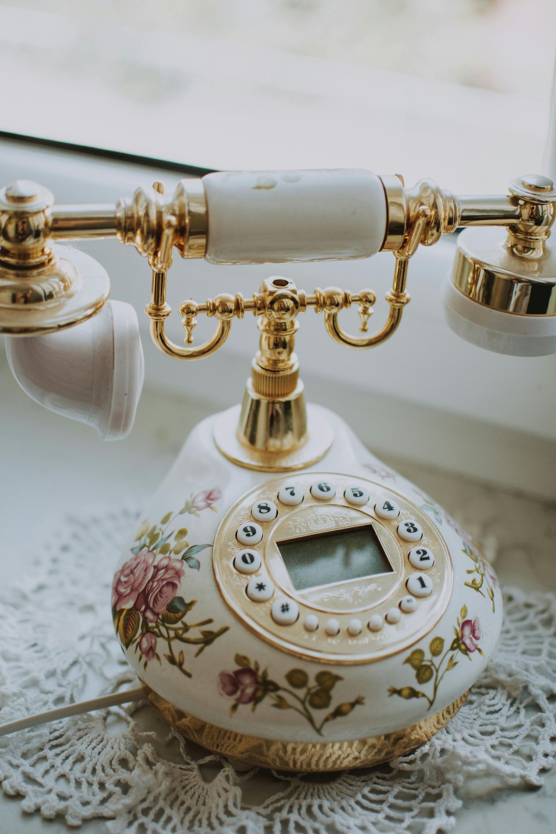 Vintage-style floral patterned phone with gold accents sitting on a white lace doily by a window.