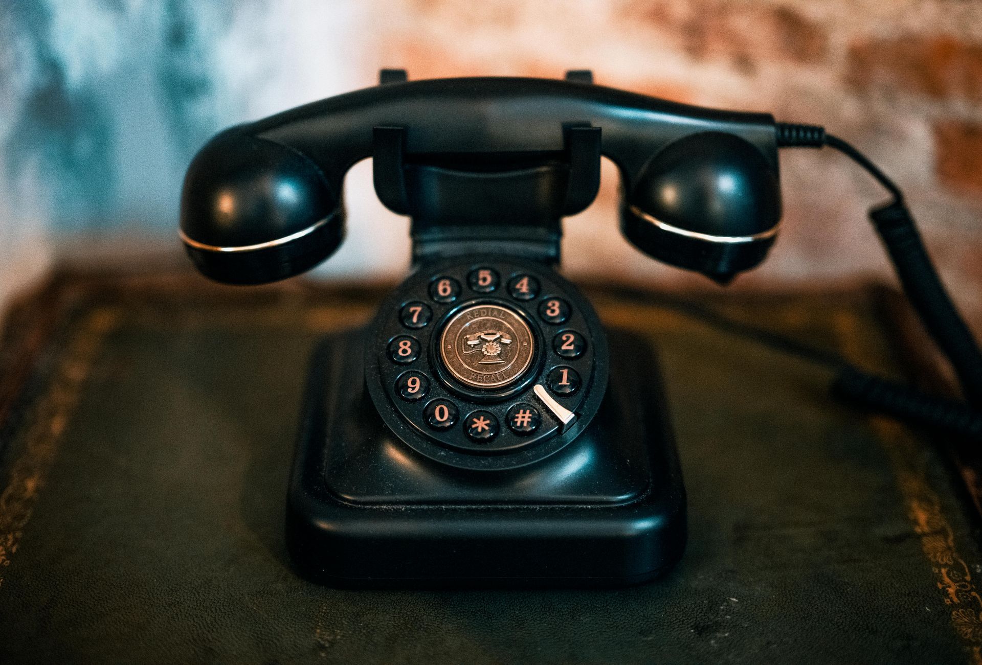 A black, vintage rotary-dial telephone sits on a textured surface against a blurred brick wall.