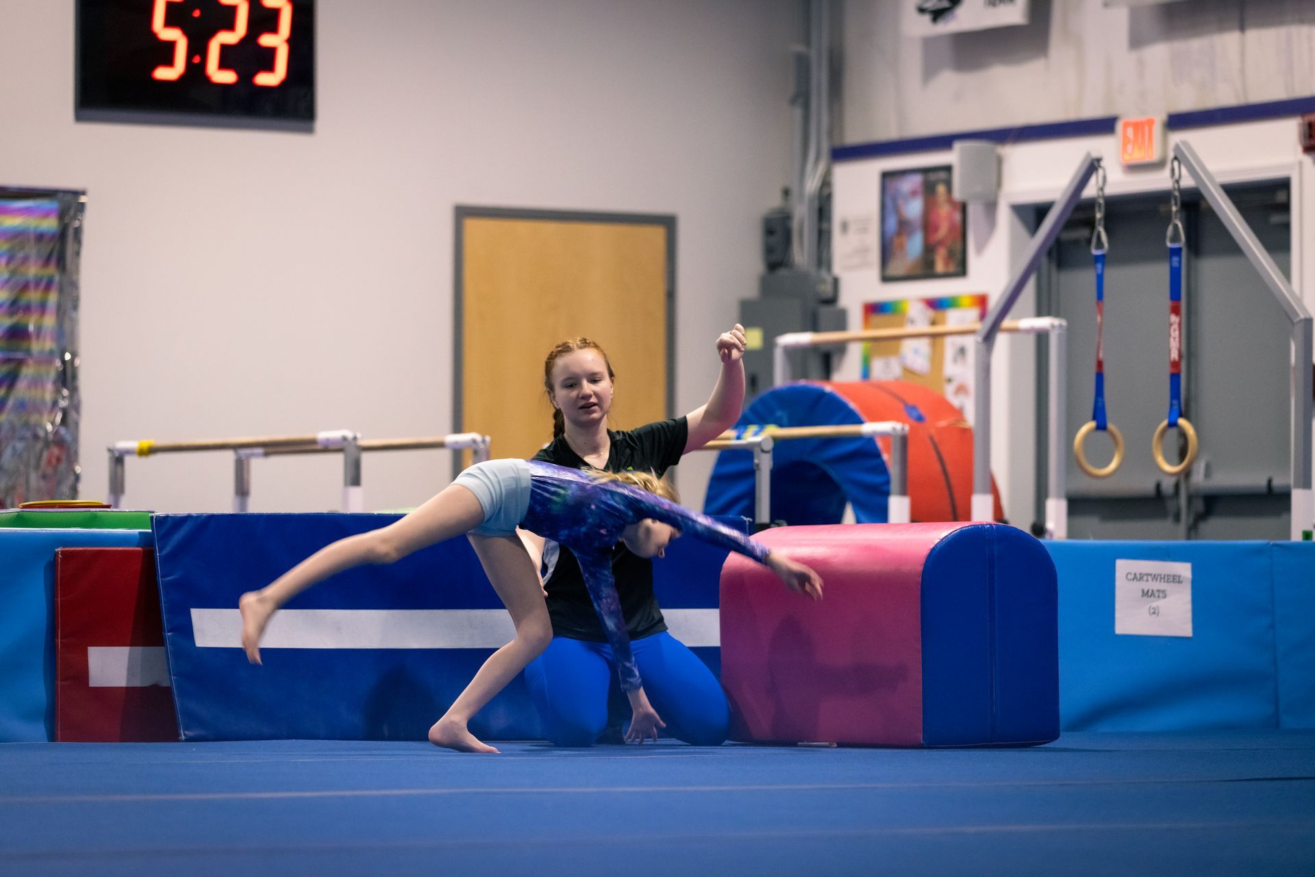 A woman is helping a young girl do a handstand on a gym floor.