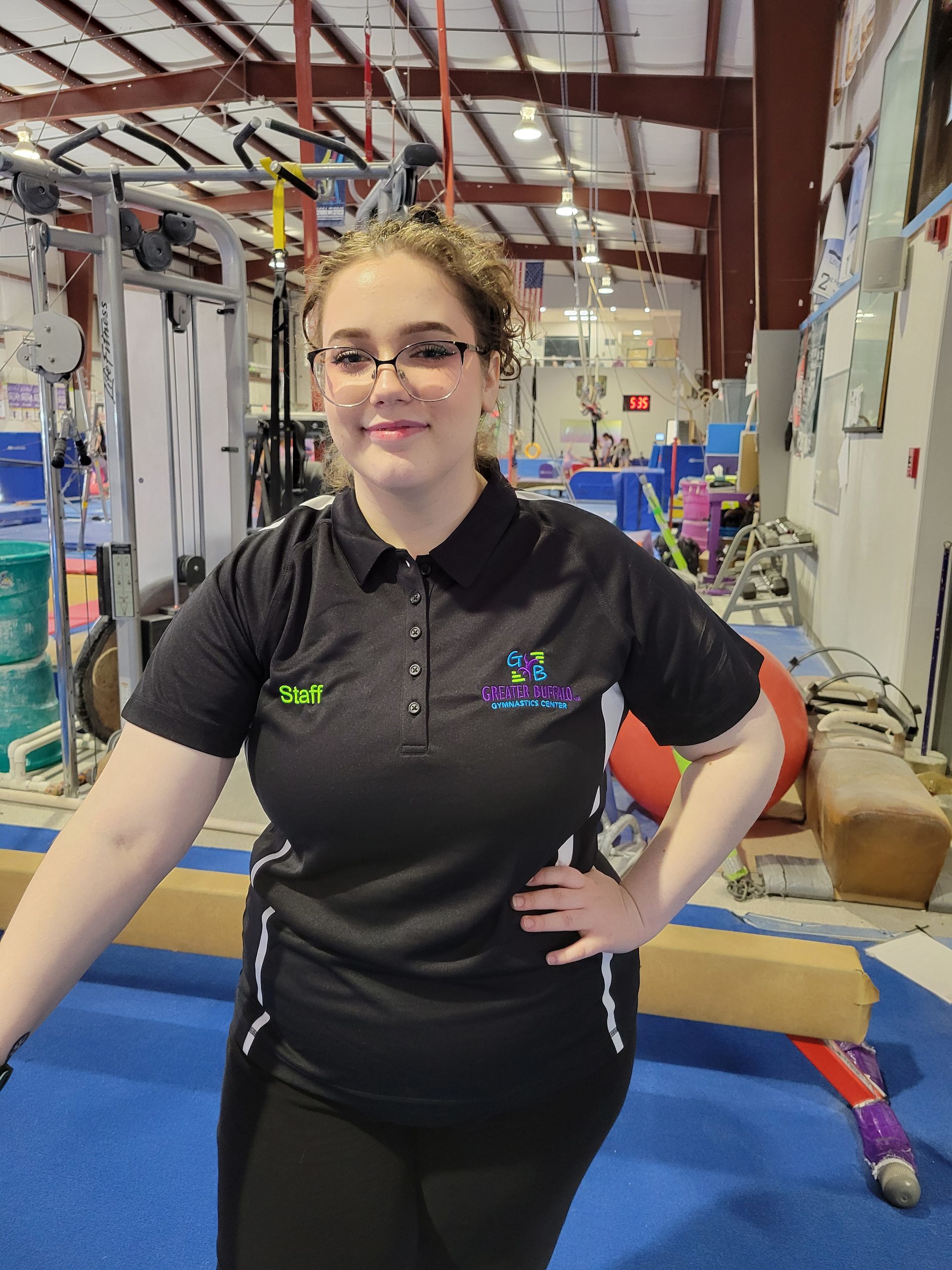 A woman wearing glasses and a black polo shirt is standing in a gym with her hands on her hips.