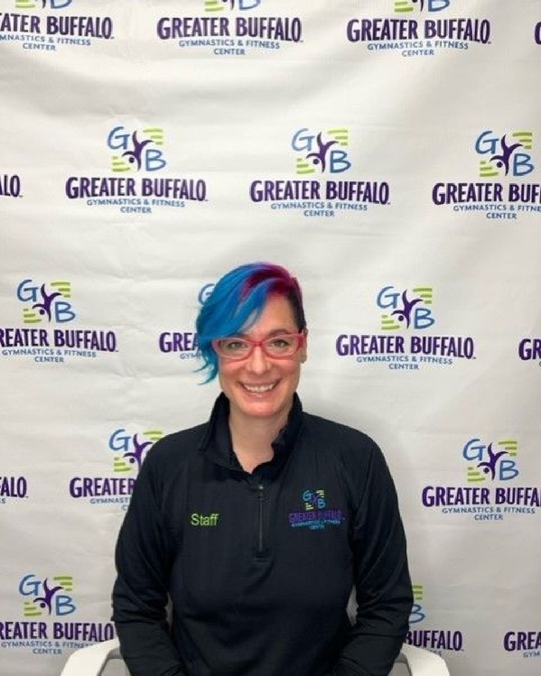 A woman with blue hair and glasses is sitting in front of a greater buffalo sign.