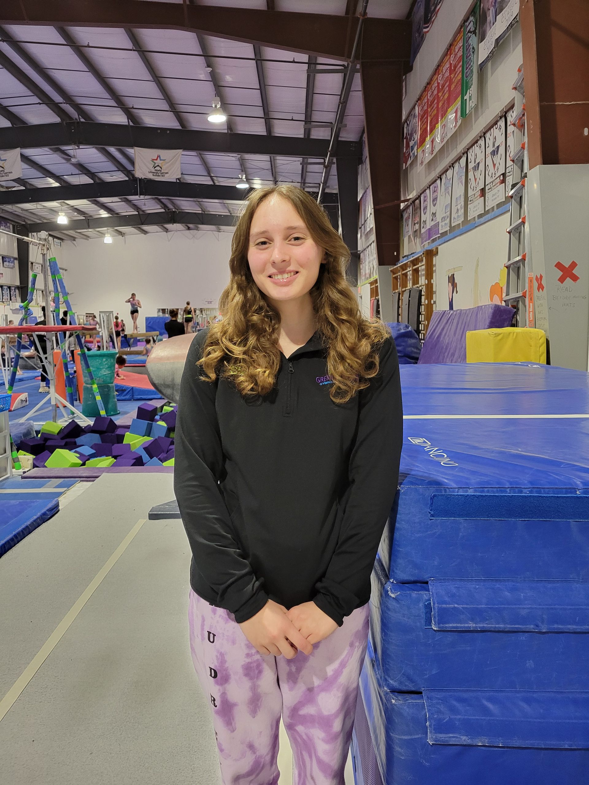 A young girl is standing in a gym next to a balance beam.