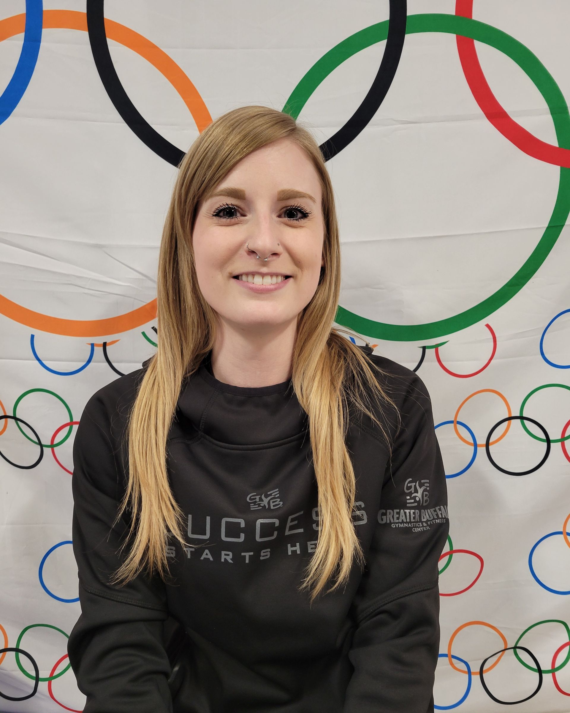 A woman is smiling in front of a wall with olympic rings on it