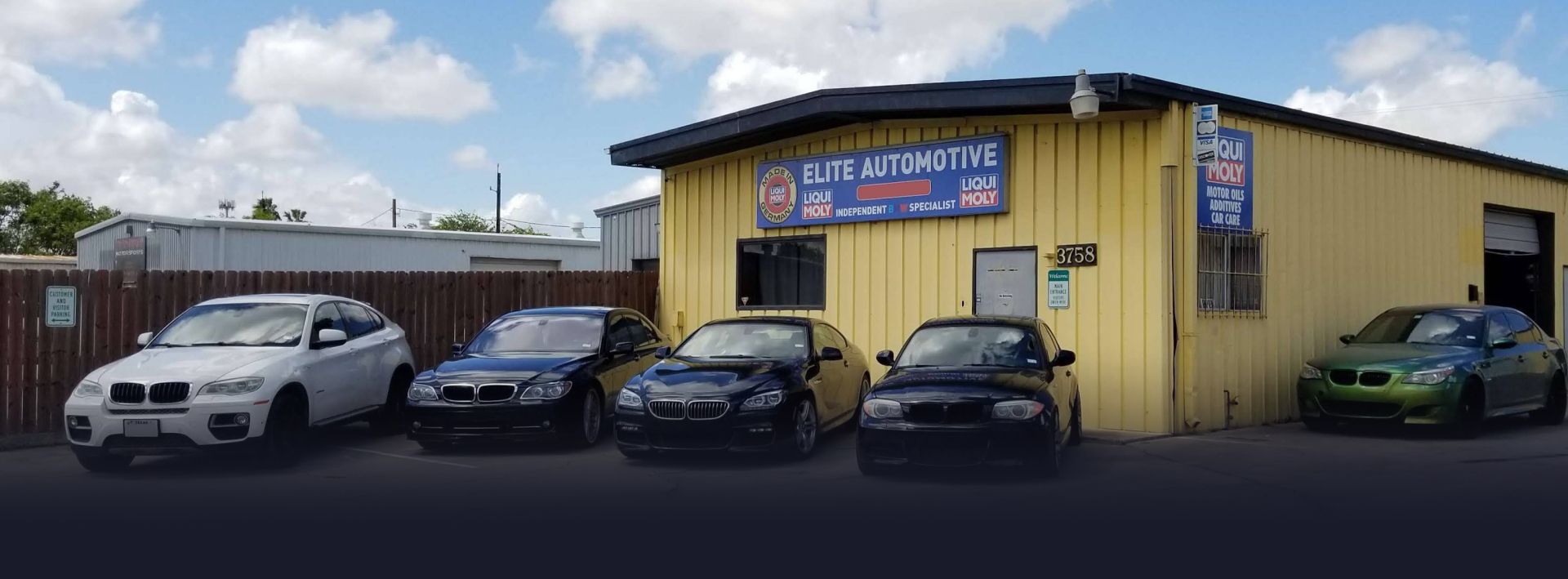 An automotive shop with five cars parked outside. Yellow building with a sign that reads, 