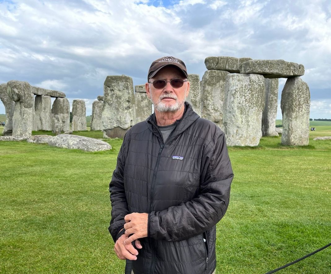 A man wearing sunglasses and a hat is standing in front of stone circles.