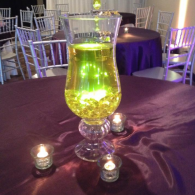 Green-lit floral vase centerpiece on a purple tablecloth, surrounded by small candleholders at an event venue