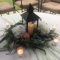 Lantern centerpiece with white flowers, candles, and greenery on a white tablecloth outdoors