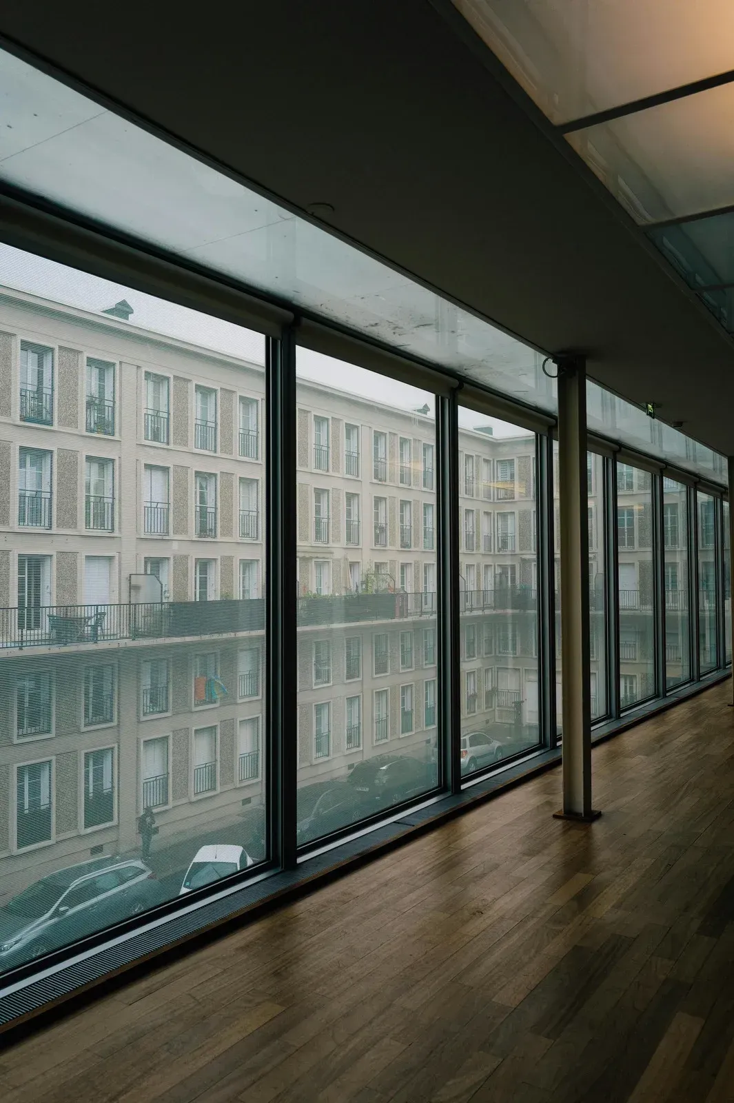 Long hallway with large windows overlooking a city street. Buildings reflect in the glass. Wooden floor.