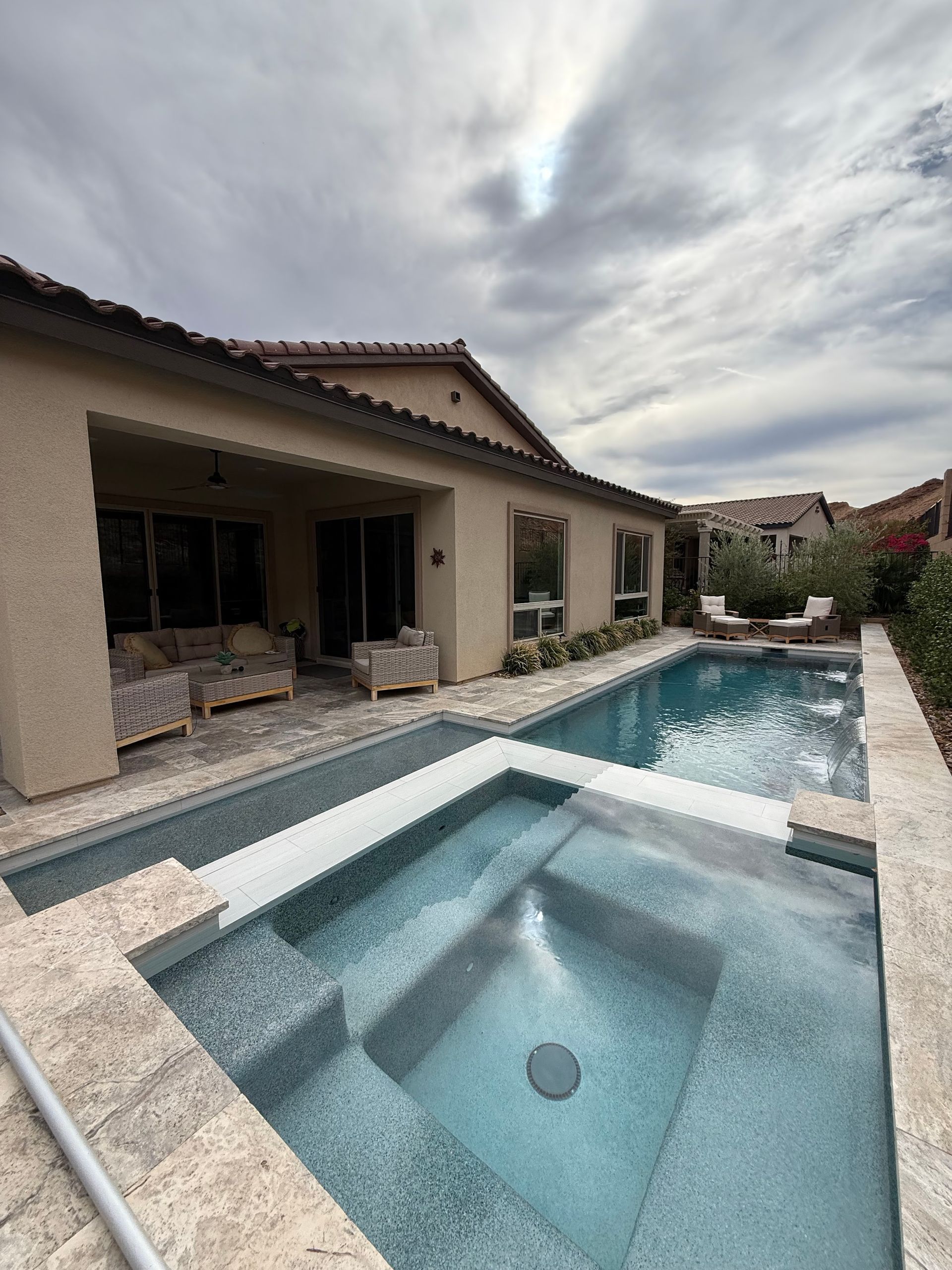Pool and spa next to a house with patio seating on an overcast day.