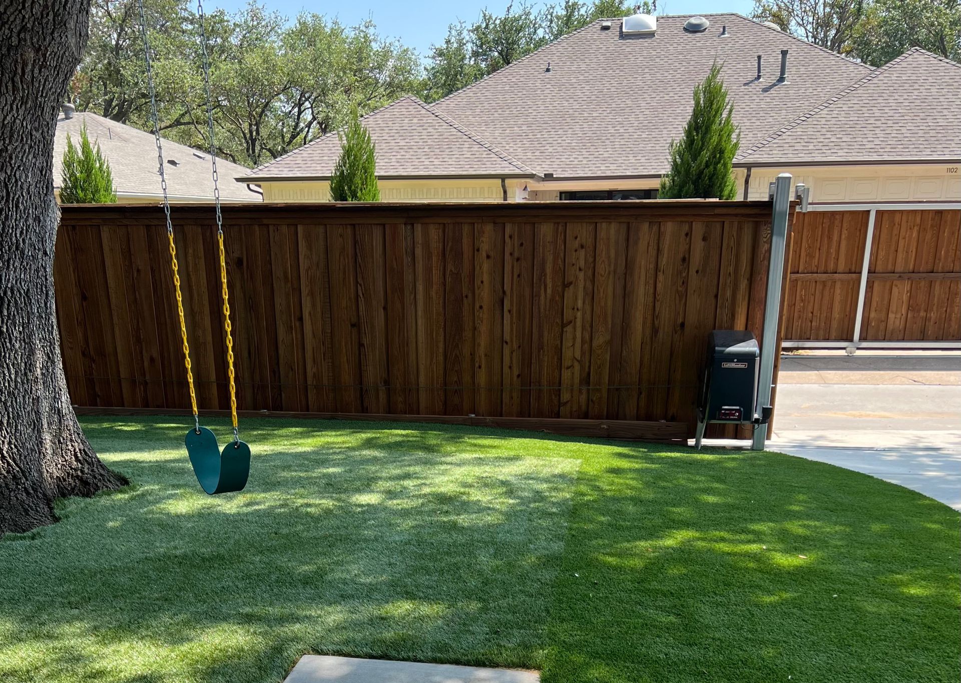 A swing is sitting under a tree in a backyard next to a wooden fence.