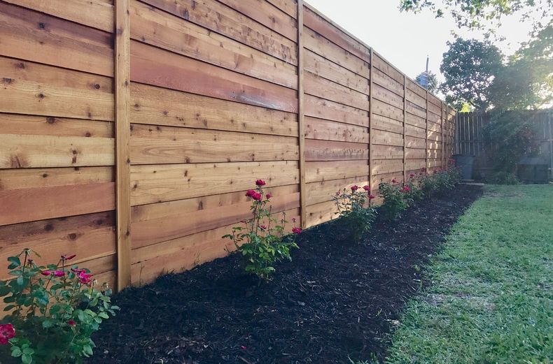 A wooden fence is surrounded by flowers and mulch in a backyard.