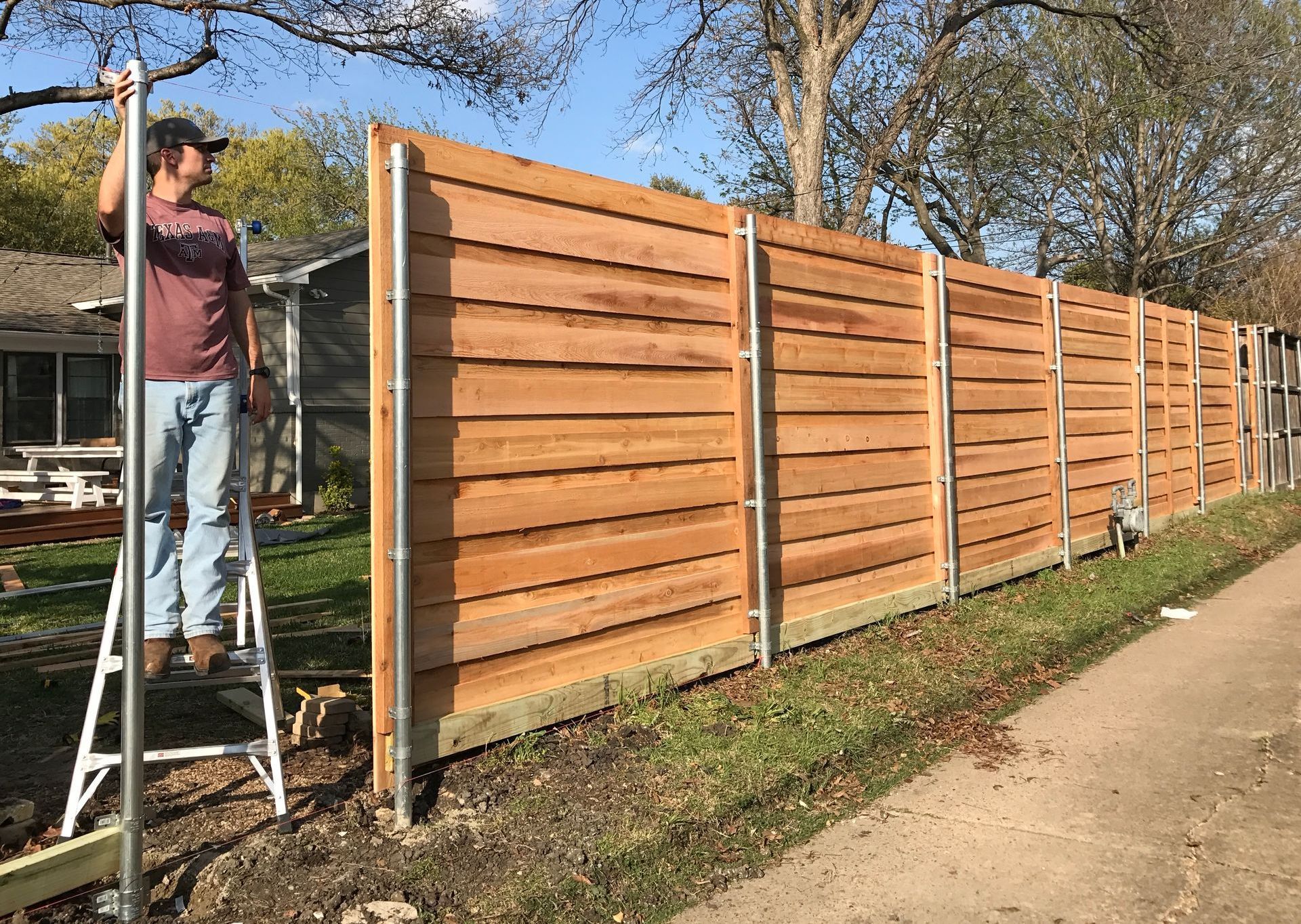 A man is standing on a ladder next to a wooden fence.