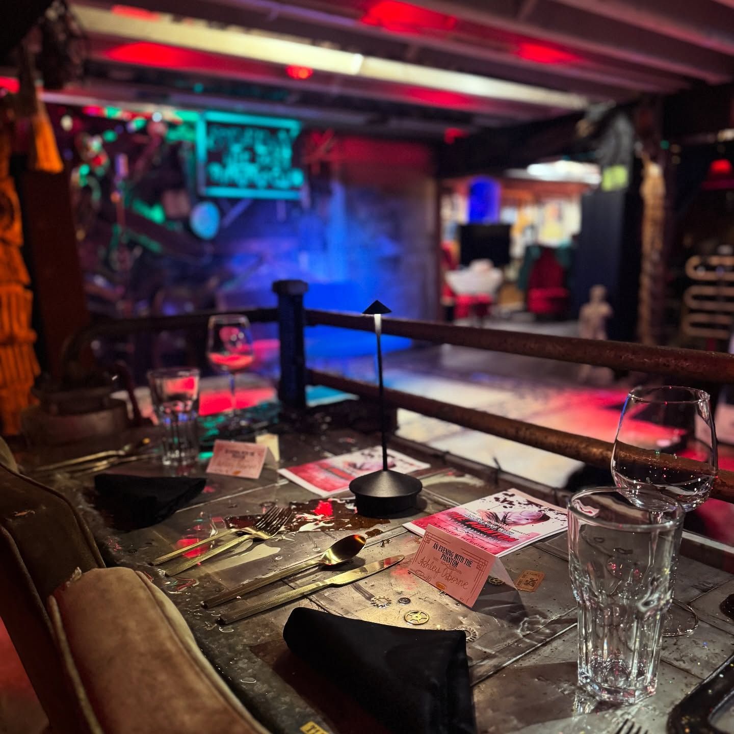 Restaurant table with glasses, cutlery, cards, and a lamp; dimly lit, red and blue lights.