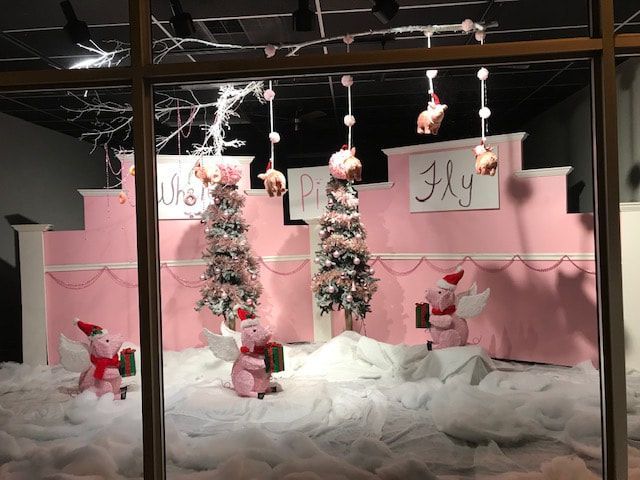Window display with pink pig figures wearing Santa hats and angel wings, snowy scene,