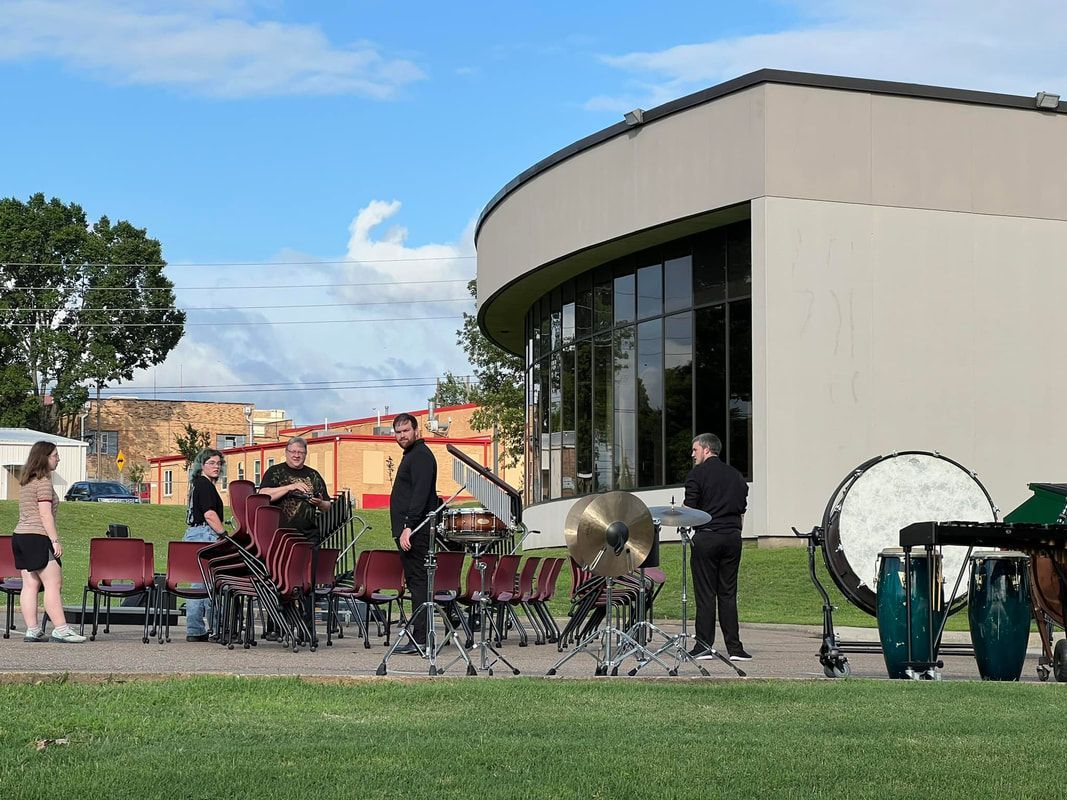 Outdoor band rehearsing; drums, cymbals, other instruments on grass near a rounded building with large windows.