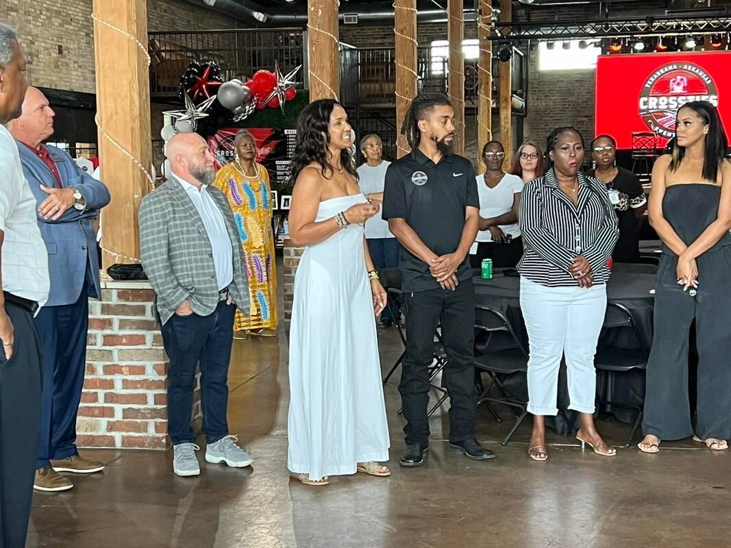 Group of people standing indoors, some in formal wear, under a red sign; brick walls and wood beams.