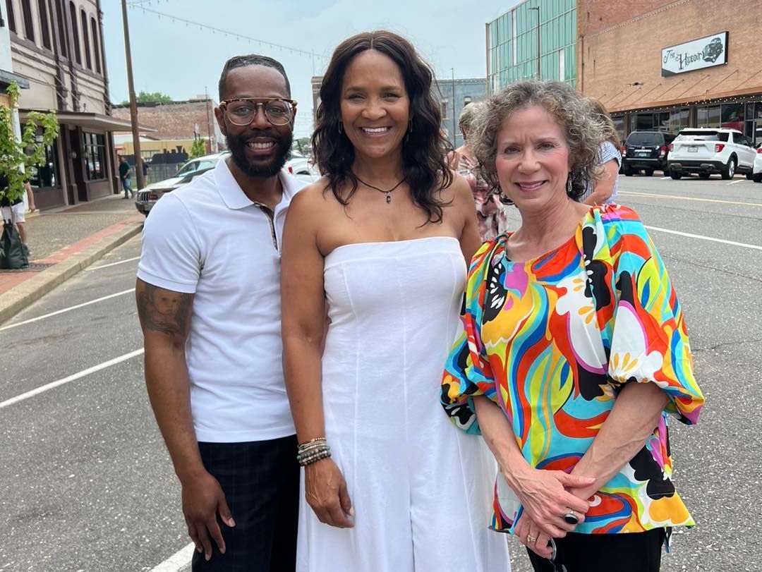Three people smiling on a city street. A woman in a white dress stands between a man in a white shirt and a woman in a colorful top.
