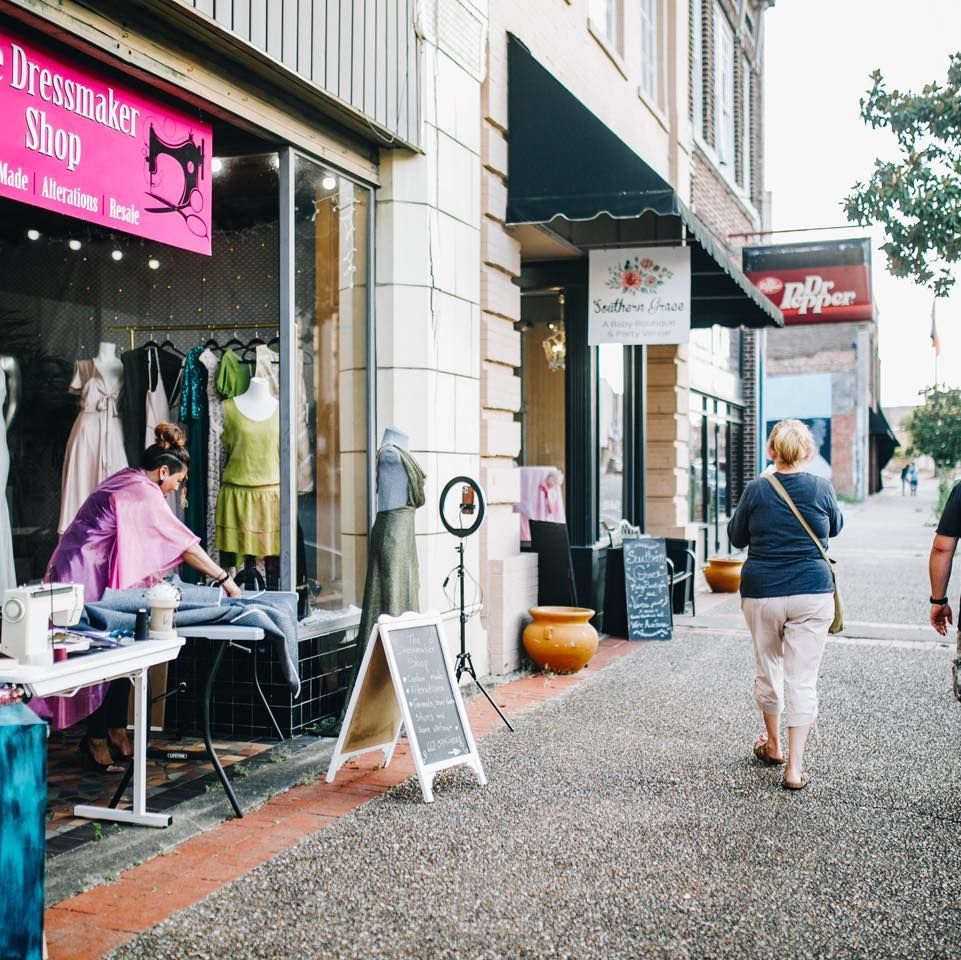 Street scene: A dressmaker works in front of shop window; two women walk on sidewalk.
