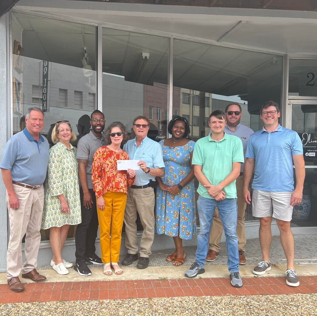 Group of people holding a check in front of a building with glass windows. Some smile.