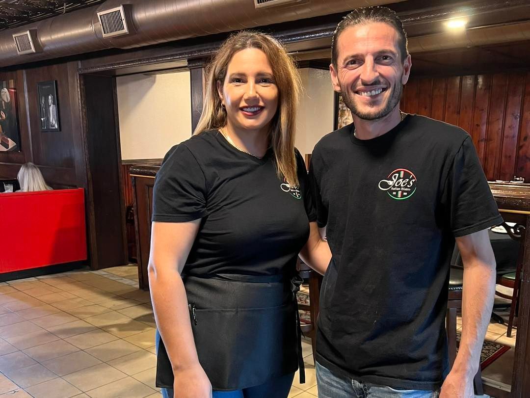Two people standing in a restaurant. Woman in black top and apron smiles. Man in black shirt smiles. Wooden interior.