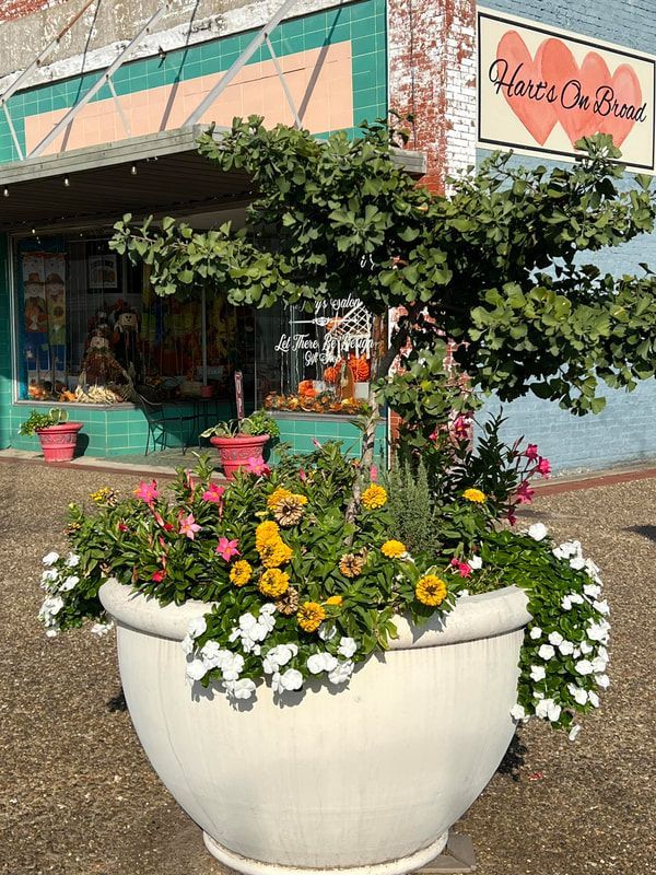 Large white planter overflowing with flowers in front of a storefront. Building has a green awning and a sign that reads 
