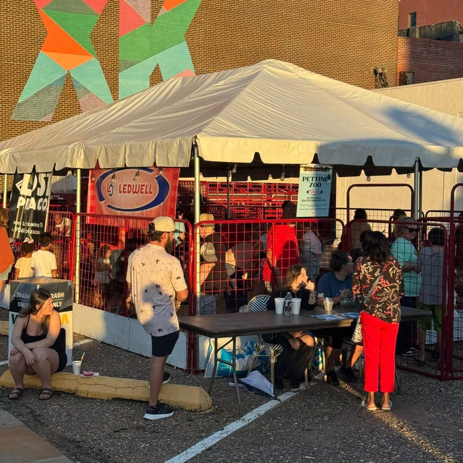 Outdoor gathering under a white tent. People standing, sitting, and interacting, with a table, red barriers, and a mural visible.