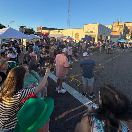 Crowd at outdoor event with tents, buildings, and a tall tower. People are gathered in a street, in the daytime.