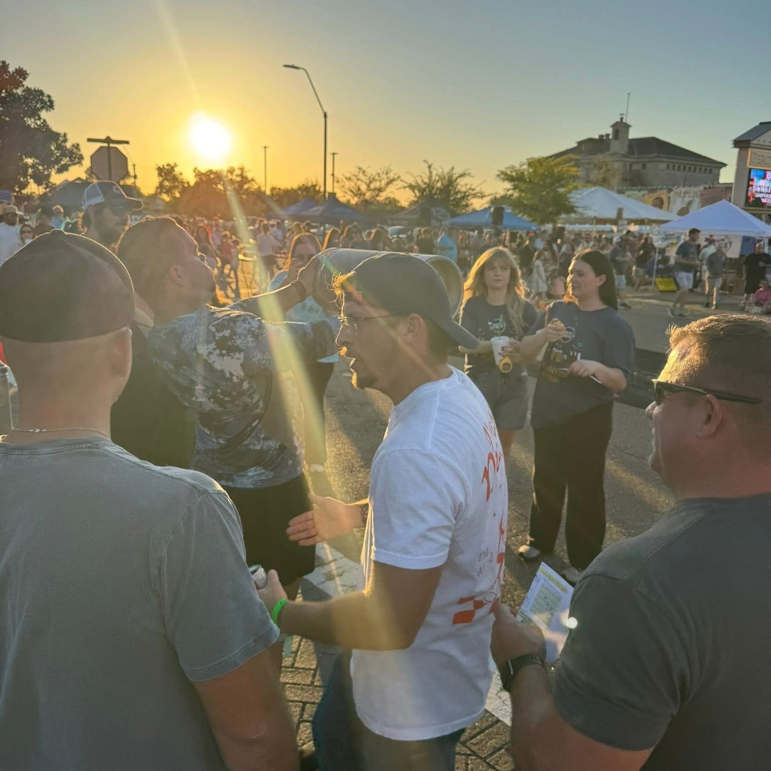 Crowd gathers outdoors at sunset, illuminated by sun. People converse and walk, in front of tents and a building.