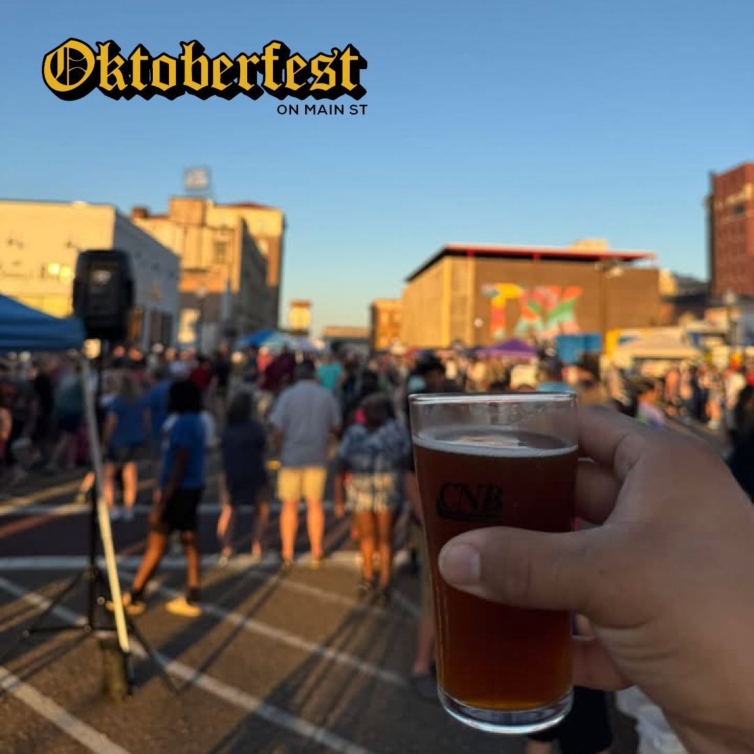 Person holding a beer at Oktoberfest on a city street; crowd in background, sunny sky.