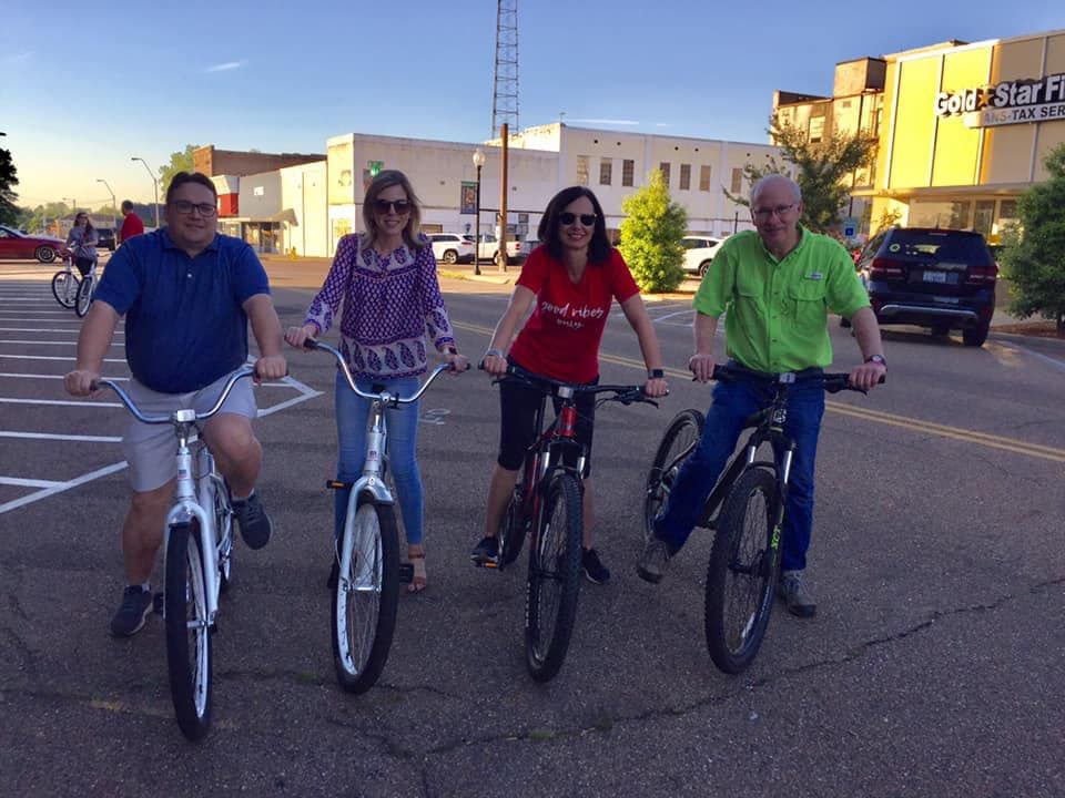 Four people on bicycles in a street. Buildings and a tower are in the background, daytime.
