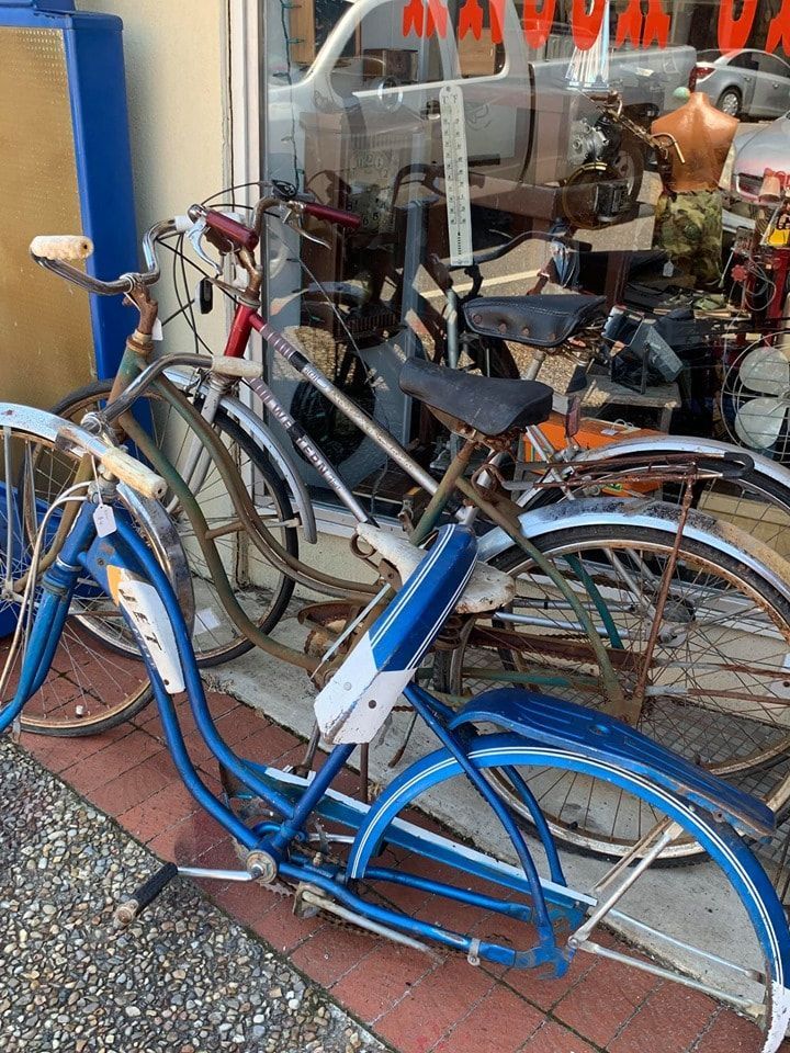 Bikes for sale leaning against a window: blue, red, and green bicycles with rusty parts.