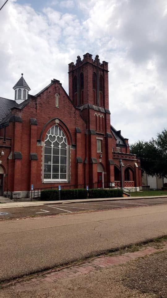 Red brick church with a tall tower, large arched window, and cloudy sky.