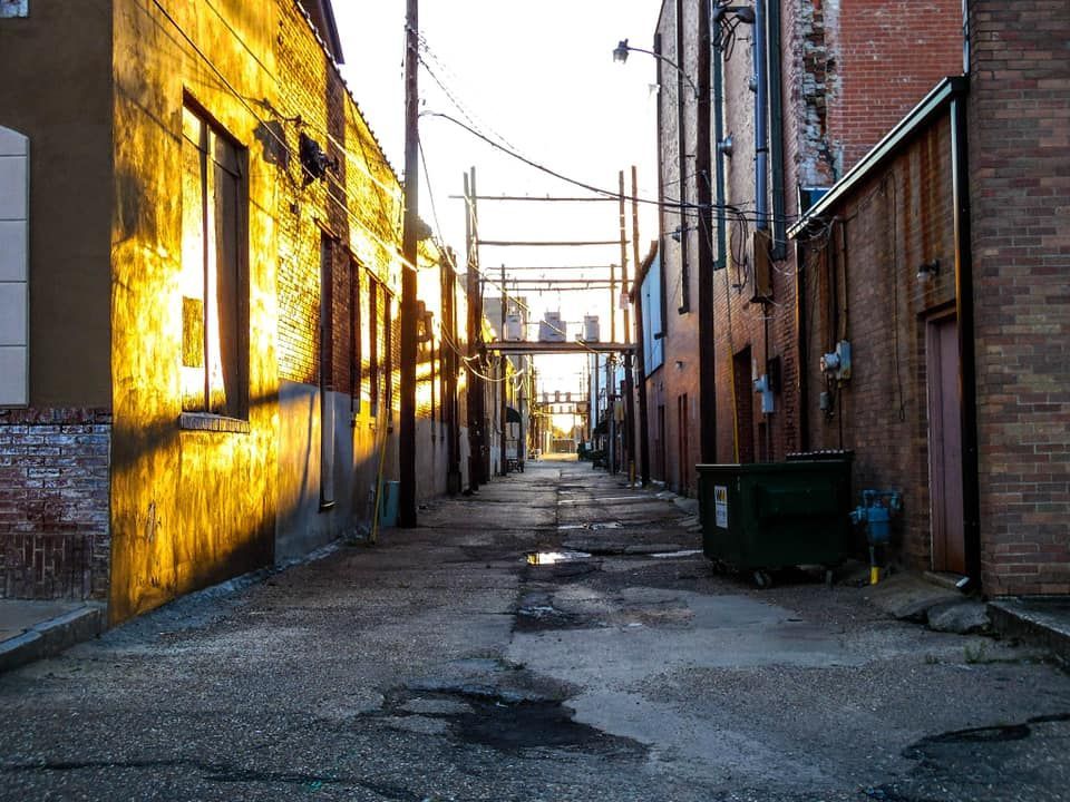 Alleyway with sunlight illuminating the left buildings. Dark, wet pavement leads to distant buildings and a metal framework.
